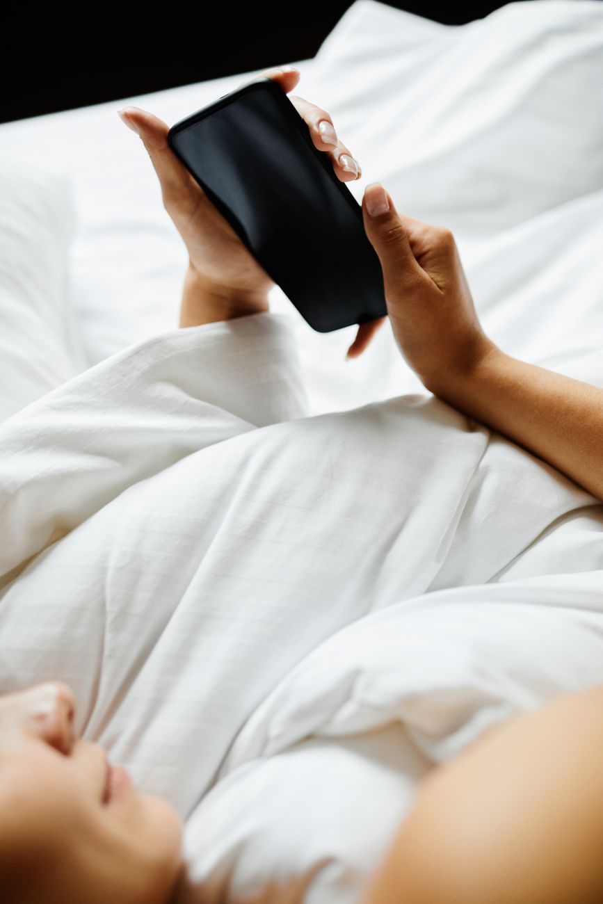A hand holding a smartphone while lying in bed under white sheets